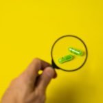 From above of crop unrecognizable male holding magnifying glass over green pills on yellow background in studio — The Precisi
