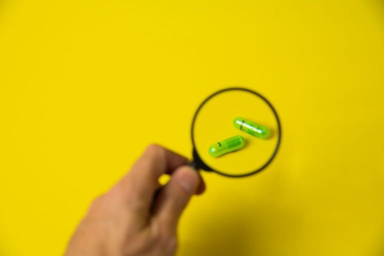 From above of crop unrecognizable male holding magnifying glass over green pills on yellow background in studio — The Precisi